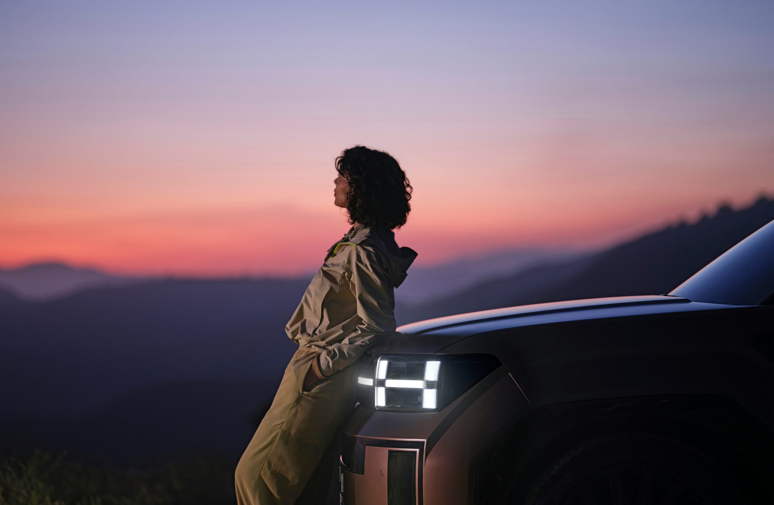 Person leaning on the front of an electric vehicle at sunset, overlooking mountain ranges in the distance.